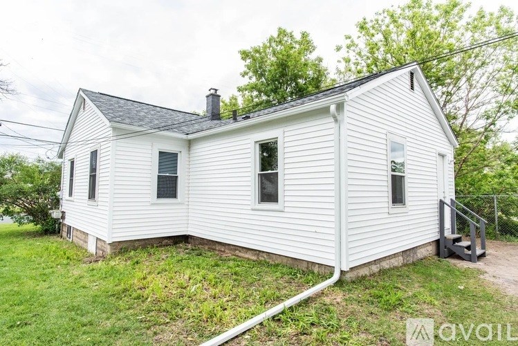 A small white house with a black roof and a chimney is surrounded by greenery.