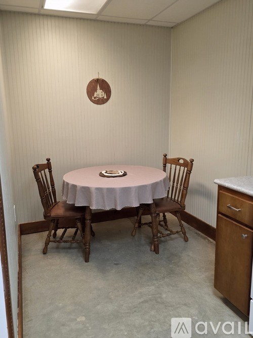 A round table with a pink tablecloth is surrounded by four wooden chairs in a room with a brown circular logo on the wall.