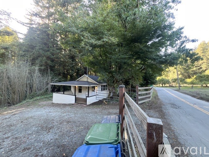 A house is situated on a gravel driveway with a fence and a green trash can.