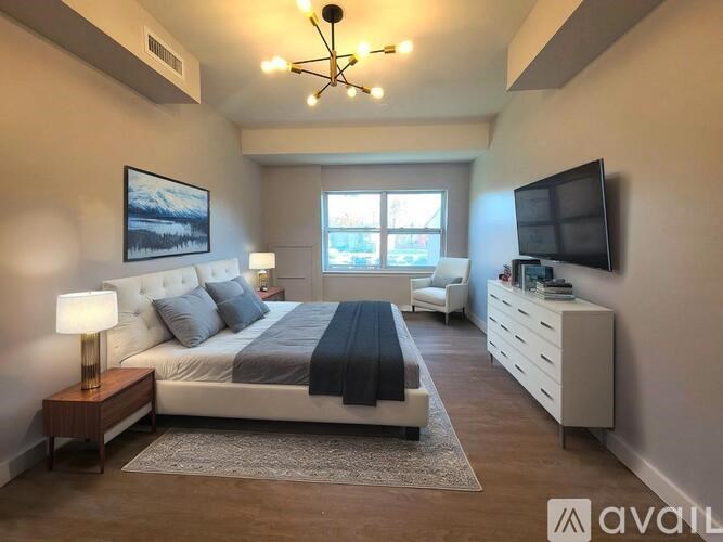 A well-lit bedroom with a large bed, a side table with a lamp, and a television mounted on the wall.