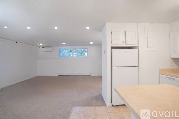 A kitchen with white appliances and cabinets, a window with a view of trees, and a beige carpet.