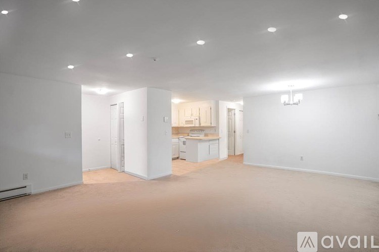 A spacious empty room with white walls and a kitchen area in the background.
