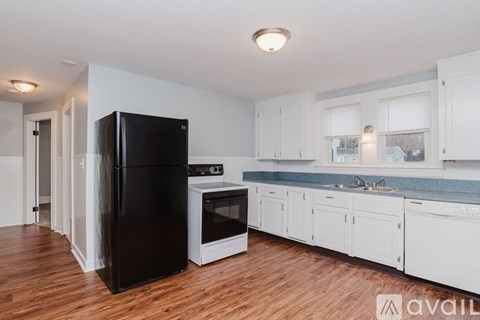 A black refrigerator stands in a kitchen with white cabinets and a wooden floor.