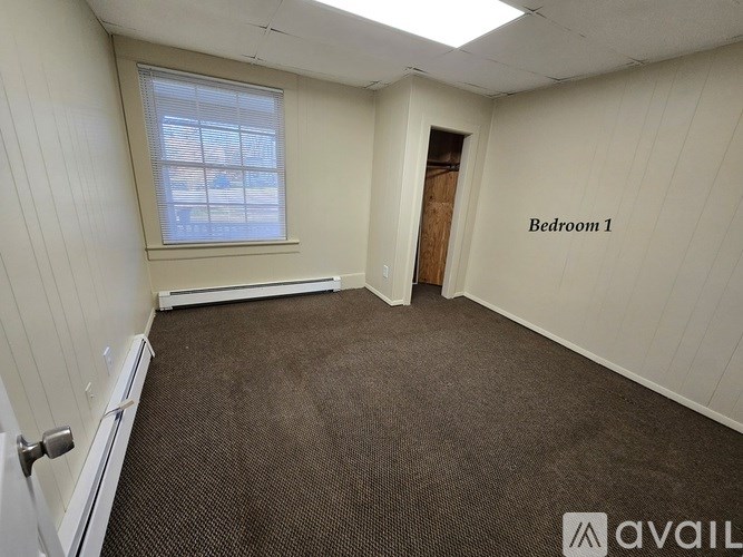 A room with a carpeted floor, a window with blinds, and a door labeled "Bedroom 1".
