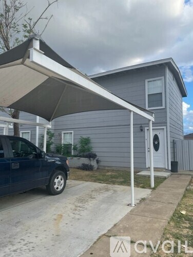A blue car is parked in front of a grey house with a white awning.