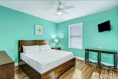 A bedroom with a bed, a dresser, a ceiling fan, a window with blinds, a wall-mounted TV, and a glass coffee table.
