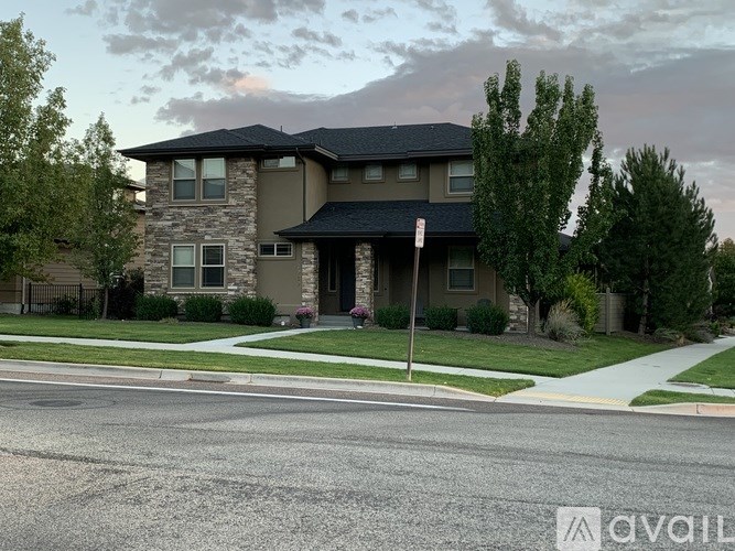 A house with a grey roof and a stone facade is surrounded by greenery and a sidewalk.