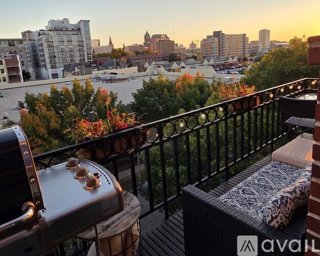A balcony with a grill and a couch overlooking a cityscape at sunset.