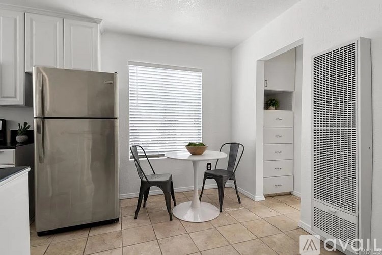 A kitchen with a refrigerator, a table with two chairs, and a cabinet with a plant on top.