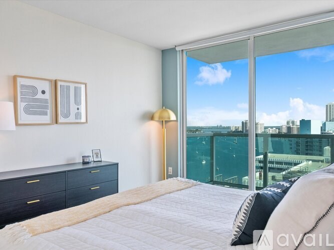 A bedroom with a large bed, a dresser, and a view of the city through a glass door.