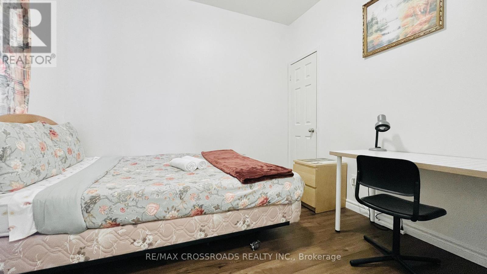 A bedroom with a floral bedspread, a desk with a chair, and a painting on the wall.