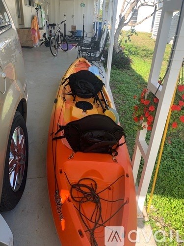 A bright orange kayak with two black bags strapped to it is on a concrete surface.