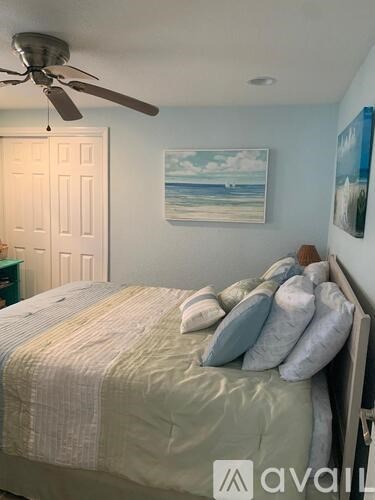 A bedroom with a bed, a ceiling fan, and a painting of a beach scene on the wall.