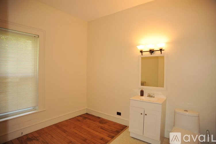 A bathroom with a wooden floor, white walls, a window with blinds, a toilet, a cabinet with a mirror above it, and a light fixture on the wall.