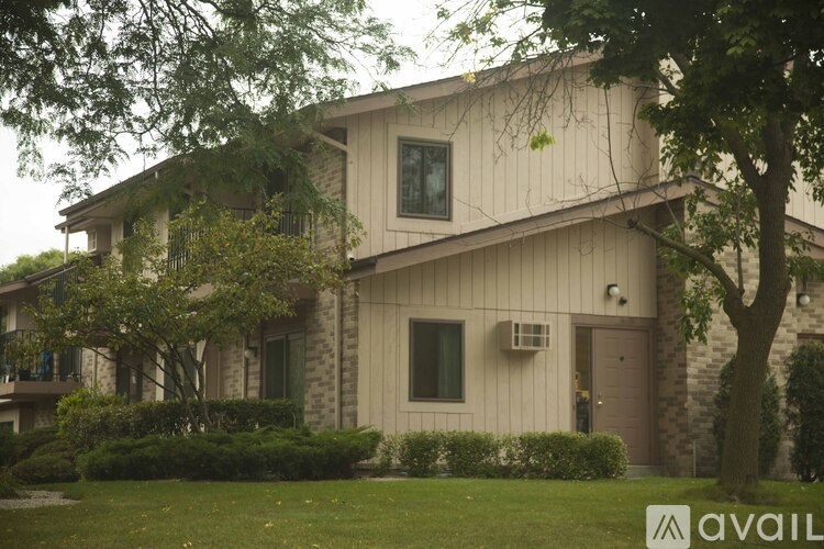 A house with a beige exterior and a brown door is surrounded by greenery.