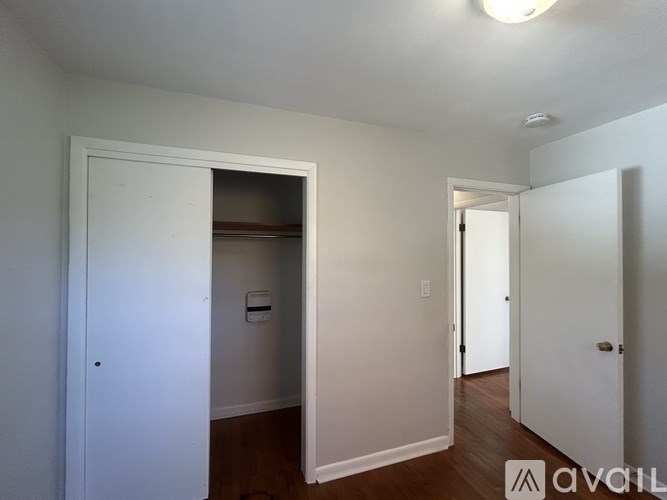 A room with white walls and wooden floors, with a door on the left and a hallway on the right.
