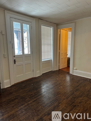 A room with wooden floors and white walls, with a door on the left and a window with blinds on the right.