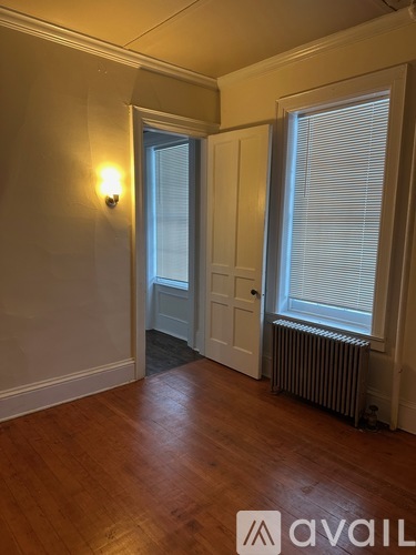 A room with a wooden floor, a window with blinds, a door, and a wall-mounted light fixture.