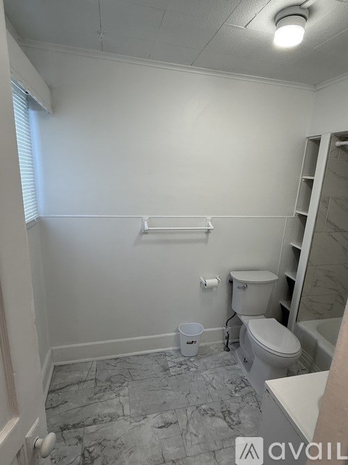 A bathroom with a toilet, a bucket on the floor, and a shelf.