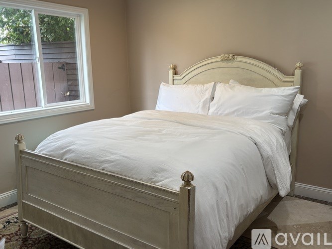 A bed with a white comforter and two pillows sits in a room with a window.