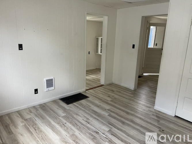 A room with wooden flooring and white walls, with a door on the right and a window in the distance.