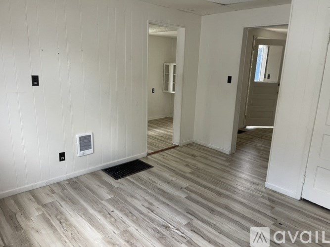 A room with wooden flooring and white walls, with a door on the right and a window in the distance.