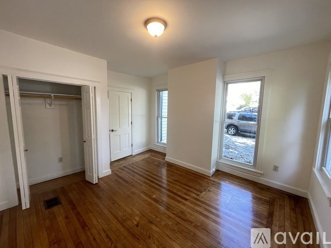 A room with wooden floors and white walls, with a window showing a parking lot outside.