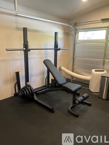 A black weight bench with a barbell on top is in a room with a window and a trash can.