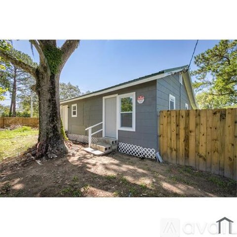 A small house with a grey exterior and a white door is surrounded by a wooden fence and a large tree.