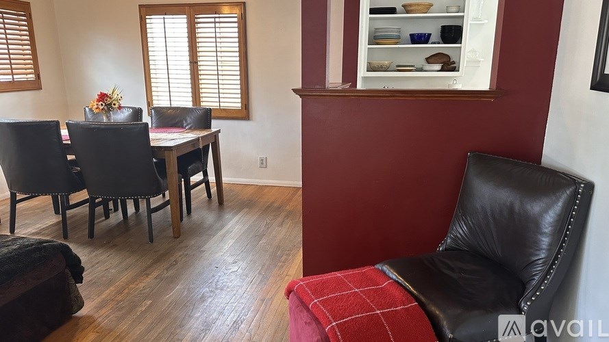 A black leather chair with a red plaid cushion sits in a room with wooden floors and a window with blinds.