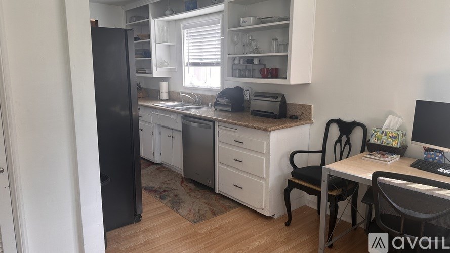 A kitchen with a black fridge, white cabinets, a desk with a computer, and a chair.