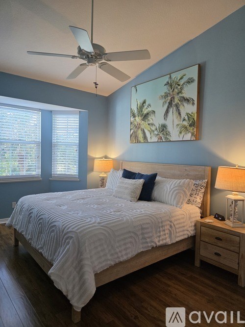 A bedroom with a bed, a ceiling fan, and a painting of palm trees on the wall.
