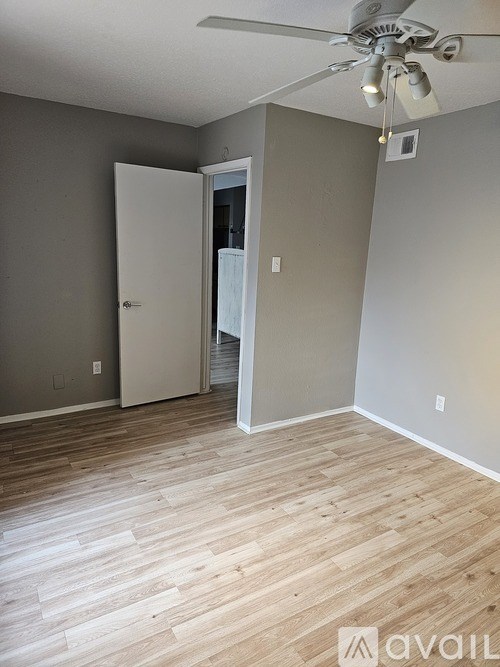 A room with a ceiling fan and light, wooden flooring, and a doorway leading to another room.