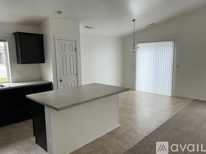 A spacious living room with a kitchen area to the left and a large window with blinds on the right.