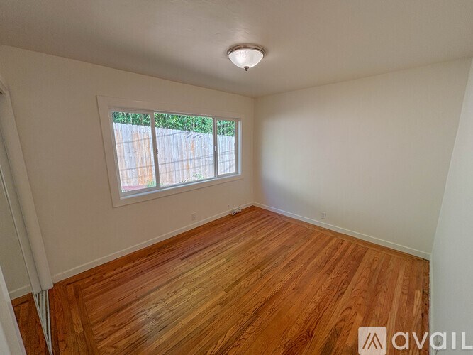 A room with wooden flooring and a window with a view of a fence and trees.