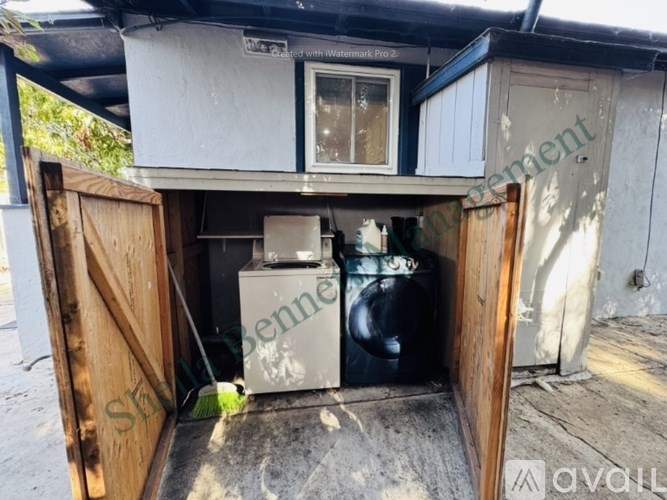 A small, dirty shed with a door open to reveal a washing machine and a black barrel.