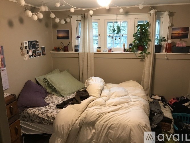 A bedroom with a bed covered in a white comforter and pillows, a window with curtains, and a string of lights hanging from the ceiling.