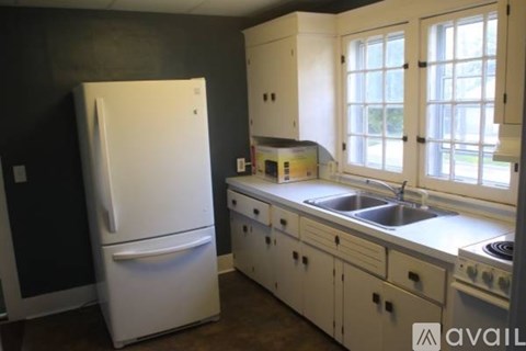 A white refrigerator stands in a kitchen with white cabinets and a window.