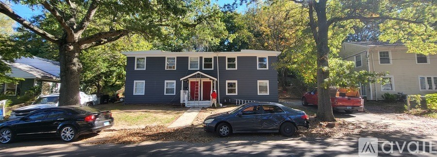 A grey house with a red door is surrounded by trees and has cars parked in front.