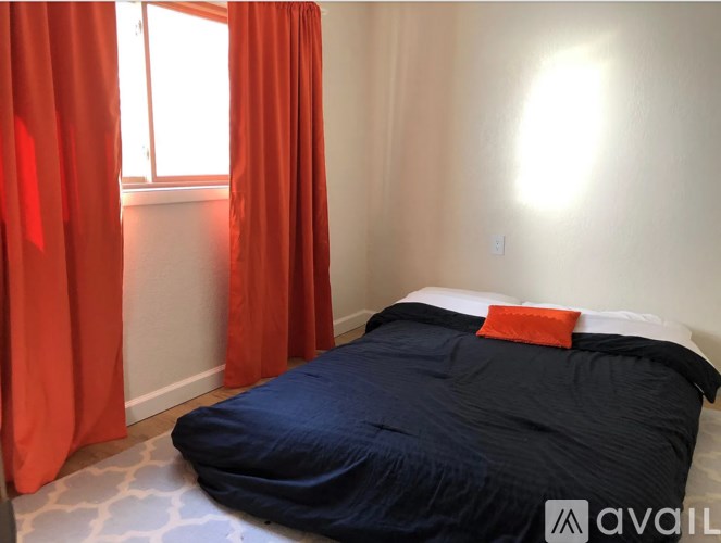 A bed with a blue comforter and an orange pillow is in a room with a window covered by red curtains.