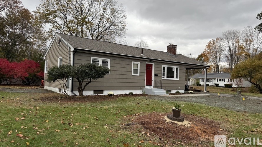 A house with a red door is surrounded by trees and has a small tree in front of it.
