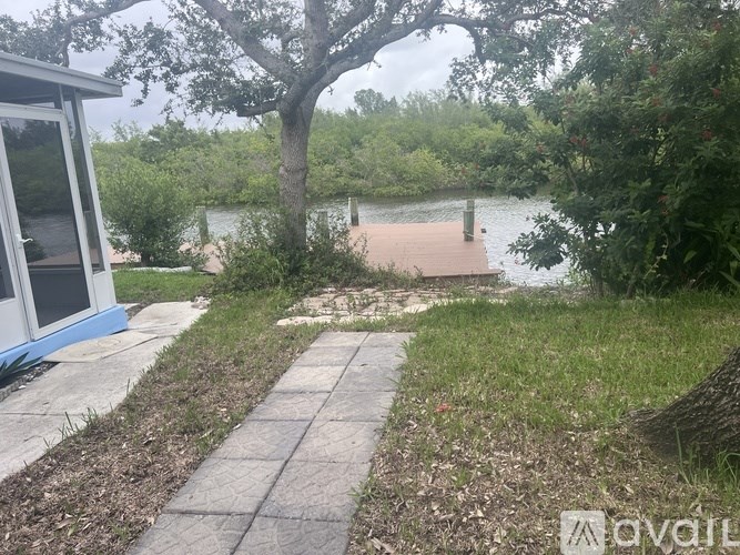 A house with a blue trim is situated on a concrete walkway next to a tree and a body of water.