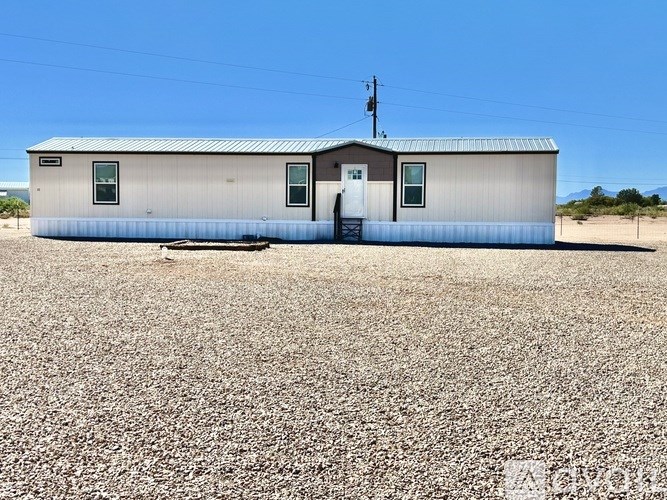 A small house with a metal roof and a door in the middle is surrounded by a gravel lot.