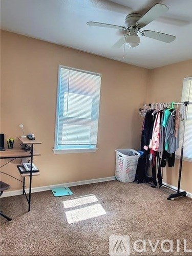 A room with a fan, a desk, a window with blinds, a laundry basket, a clothes rack with clothes, and a green mat on the floor.