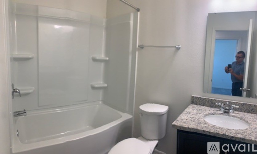 A man is taking a picture of a bathroom with a white toilet, a sink, and a bathtub.