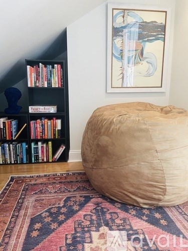 A large round pillow is on a rug in front of a bookshelf and a painting.