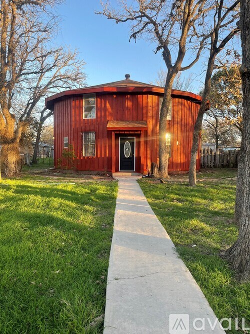 A red wooden house with a black door and windows is surrounded by trees and grass.