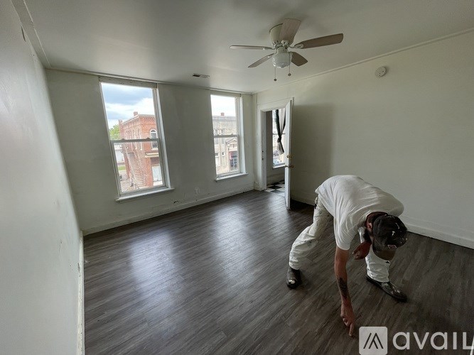 A man in a white shirt and white pants is bending over in a room with a ceiling fan and large windows.