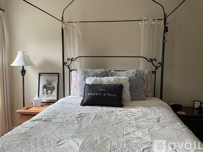 A bed with a grey comforter and a black pillow with the text "my happy place" on it.