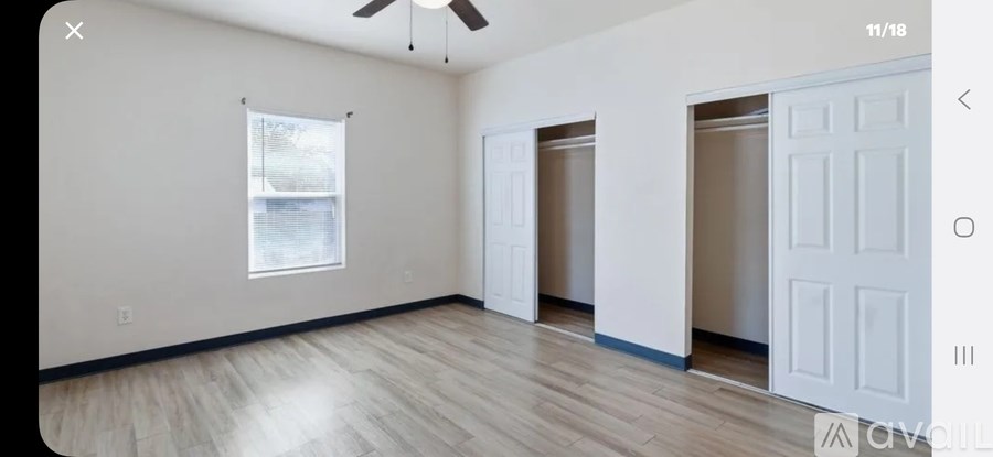 A room with white walls and wooden flooring, featuring a window with blinds, a white door, and a ceiling fan.